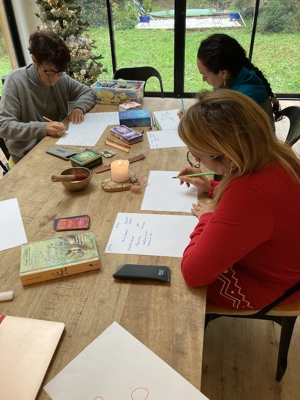 Trois participantes assises autour d'une table, en train de cartographier leurs liens familiaux au crayon sur de grandes feuilles. Bougie, bol chantant et cartes posés au centre.