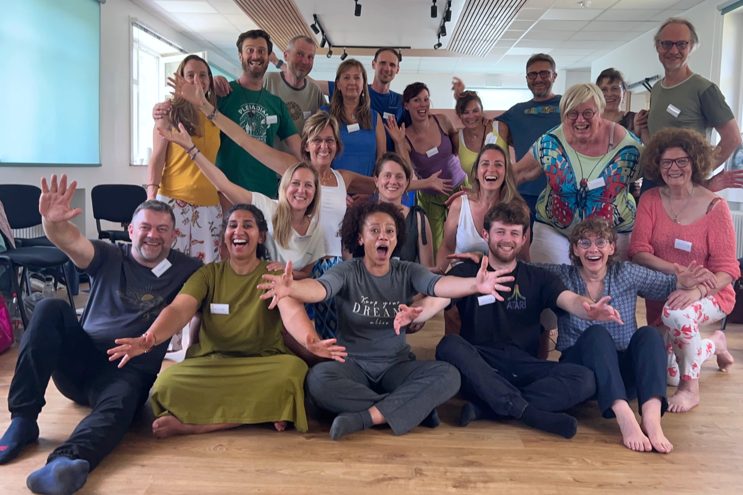 Le groupe d'une session NSQ® en 2025 : une vingtaine de participants pieds nus dans une salle de formation, assis au sol et debout derrière, bras tendus vers l'objectif, en train de rire.